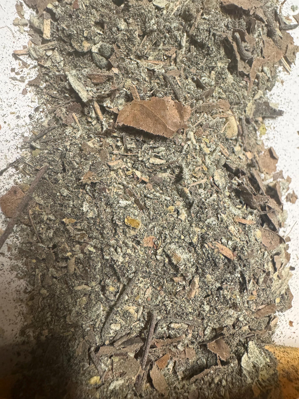 Close-up of a pile of CrewCrumble. The mix includes various shades of brown, green, and tan, with visible pieces of leaves, stems, and small granules. The texture appears coarse and natural.